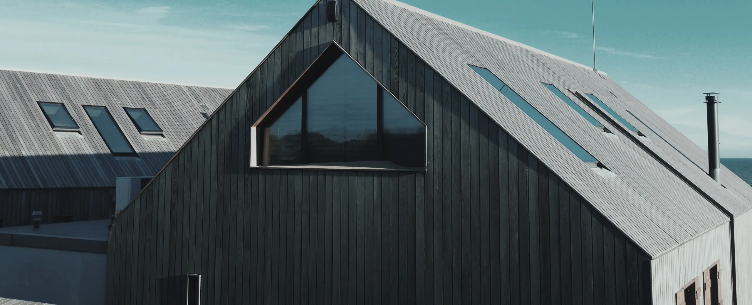 A grey wooden house with a sloping roof situated near a calm body of water, surrounded by lush greenery and trees under a clear blue sky. The tranquil scene reflects the house's image in the water.